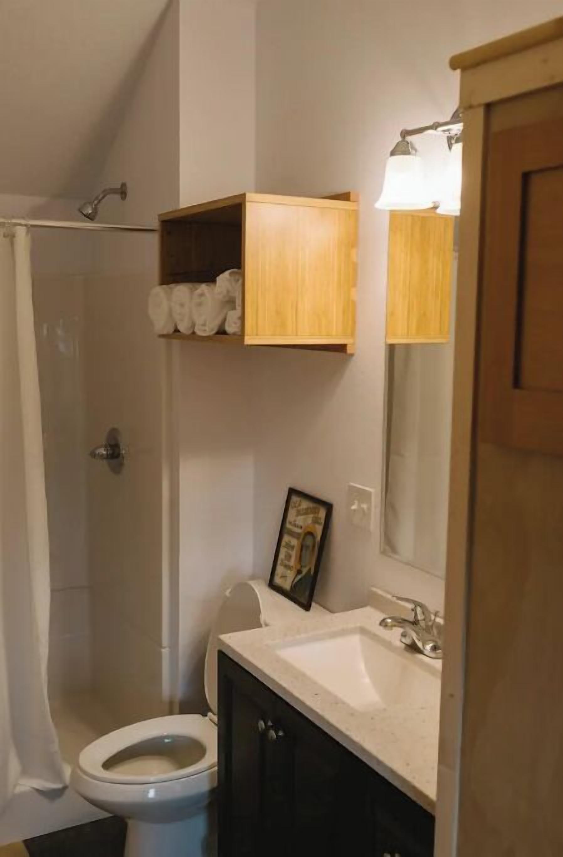 A bathroom with a shower, toilet, sink, and a wooden shelf above the toilet.