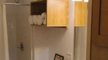 A bathroom with a shower, toilet, sink, and a wooden shelf above the toilet.