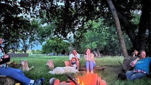 A group of people sitting around a campfire in a grassy outdoor area with trees in the background. - Flinthills Premier Getaway (Severy)