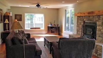 A living room with a stone fireplace, a ceiling fan, and a view of a deck.