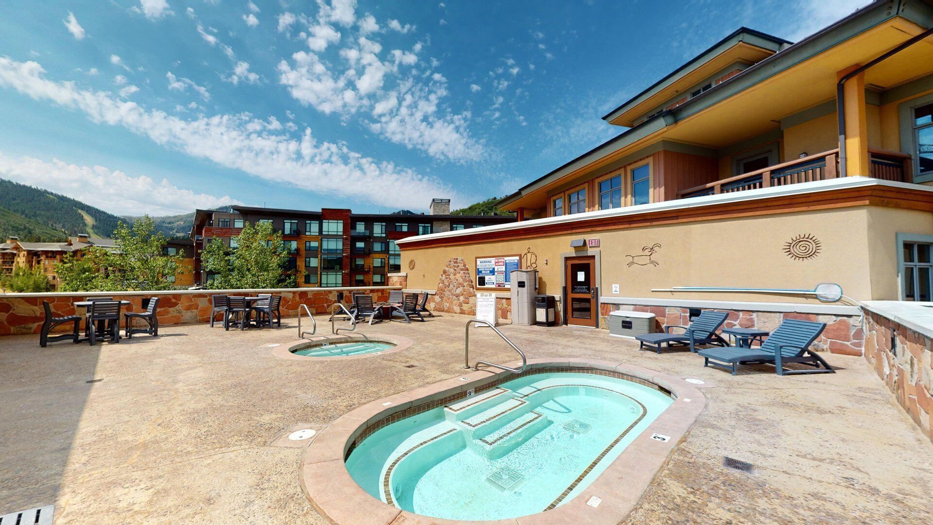 A rooftop pool area with lounge chairs and a hot tub, surrounded by a building with a stone facade and a view of mountains.
