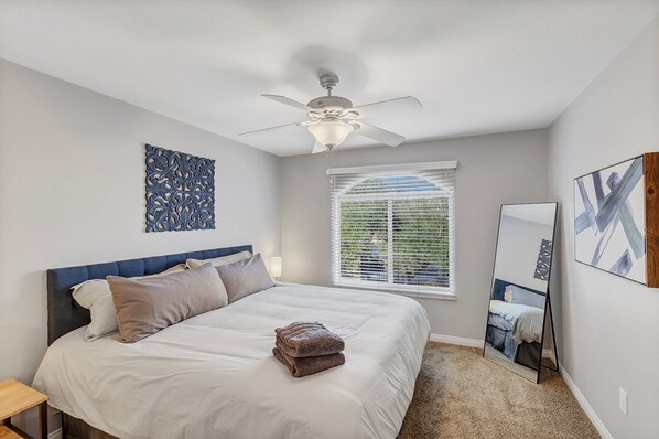 Une chambre à coucher comprenant un lit, un ventilateur de plafond, une fenêtre avec des stores, un miroir fixé au mur et un élément décoratif mural.