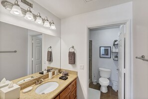 Une salle de bain équipée d’un lavabo, d’un miroir, d’un porte-serviettes et de toilettes visibles à travers une porte ouverte.