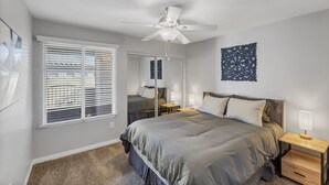Une chambre à coucher comprenant un lit, des tables de chevet, un ventilateur de plafond, une fenêtre avec des stores et un élément de décoration murale.