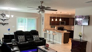 A modern living room with a black leather sofa, a coffee table, and a ceiling fan. A kitchen with dark cabinets and a TV is visible in the background. - Brand New Construction 🏠 Resort style Getaway (Jacksonville)