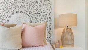 Ein ordentlich bezogenes Bett mit einer gestreiften Tagesdecke und mehreren Kissen, ein dekorativer Wandvorhang als Kopfteil, eine Nachttischlampe und ein kleiner Tisch mit einer Vase und einer Blume.