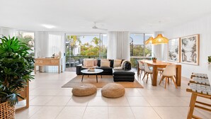 Ein geräumiger Wohnbereich mit einer Couch, Poufs, einem Esstisch und Stühlen. Ein Balkon bietet Blick auf Palmen.