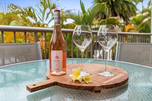Una botella de vino rosado, dos copas de vino vacías y una tabla de madera sobre una mesa de vidrio, con vistas tropicales al fondo.