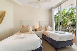 Habitación de hotel con dos camas, ventilador de techo, balcón con vista a palmeras y un gran ventanal.