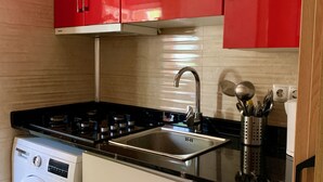 Une cuisine de taille réduite avec des meubles rouges, un évier en acier inoxydable, un lave-linge et un plan de travail noir.