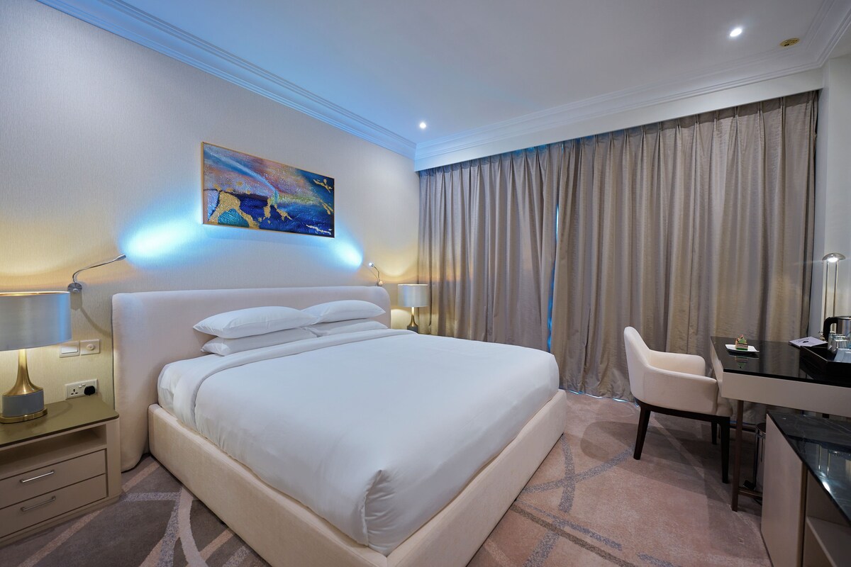 Une chambre d’hôtel avec un grand lit, un bureau et une chaise. Il y a un tableau au mur et une lampe sur la table de chevet.