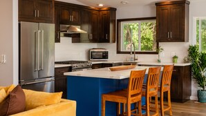 Cocina moderna con armarios de madera oscura, electrodomésticos de acero inoxidable, una isla azul con taburetes y vistas a zonas verdes.