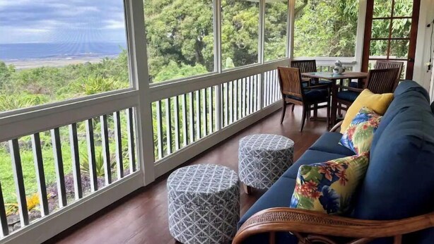 Un porche cubierto con vistas a un paisaje exuberante, un sofá azul con cojines estampados y una mesa de comedor con sillas.