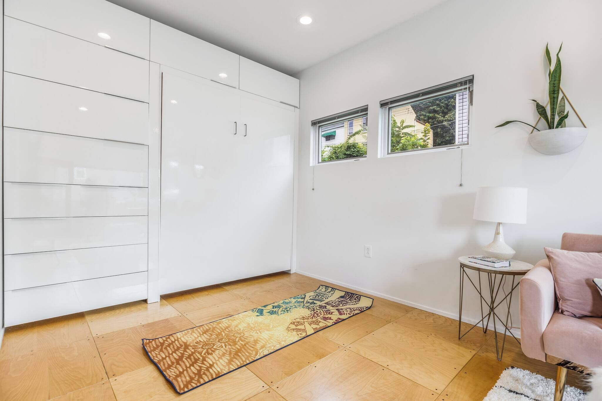 A modern kitchen with white cabinets, a wooden floor, and a patterned rug. There are two windows with blinds, a small side table with a lamp, and a potted plant on the wall.