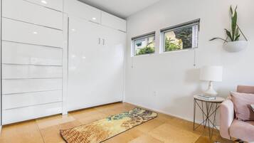 A modern kitchen with white cabinets, a wooden floor, and a patterned rug. There are two windows with blinds, a small side table with a lamp, and a potted plant on the wall.