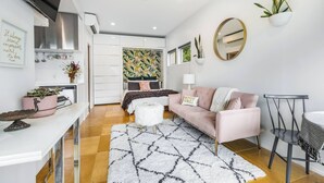 A modern living room with a white sofa, a coffee table, and a patterned rug. There is a kitchen area with white cabinets and a stainless steel range hood. - Not so tiny house with full kitchen & patio swing (Pittsburgh)