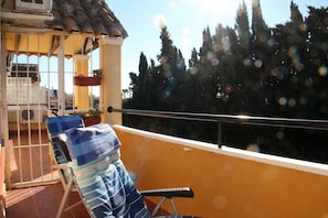 A balcony with a chair and a small table, overlooking a garden and tall trees.