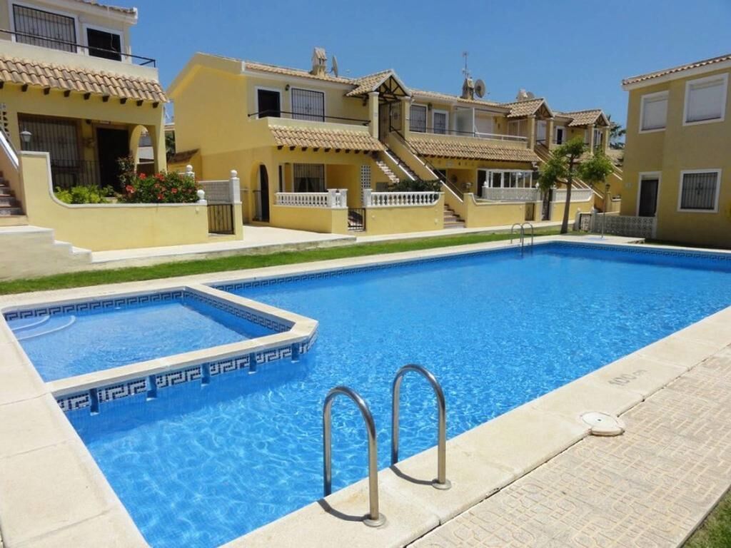 A swimming pool with a tiled deck, surrounded by yellow buildings with balconies and a clear blue sky.