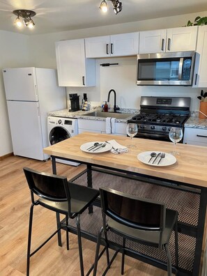 A compact kitchen with a microwave, stove, sink, and a washing machine. There is a wooden dining table with chairs and a small island with a sink.