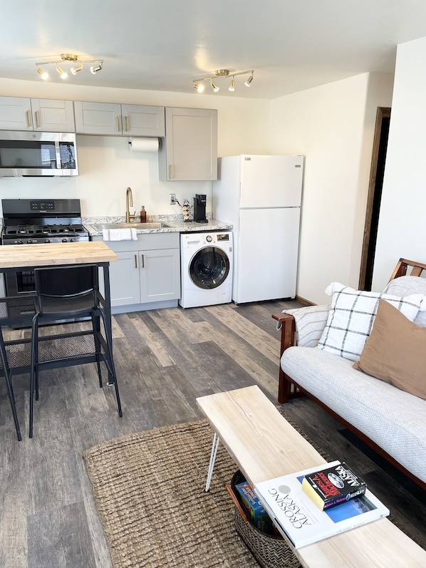 Un espacio habitable compacto con kitchenette, zona de comedor y área de estar con sofá y mesa de centro.