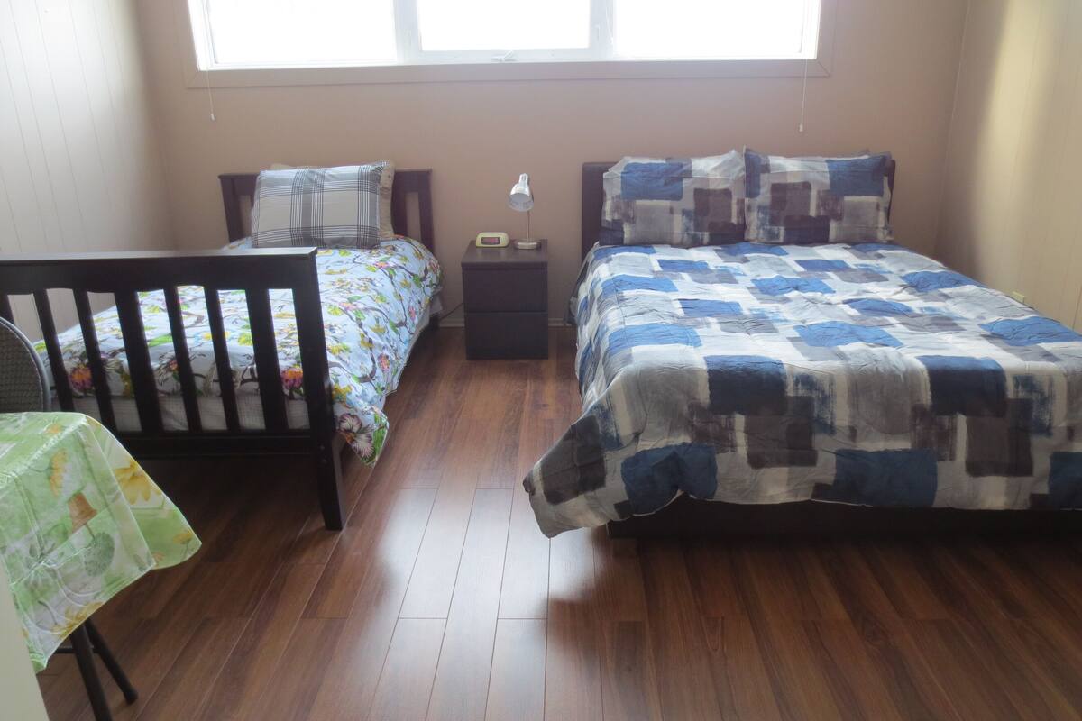 Quarto com piso de madeira, berço, cama com colcha azul e branca e mesa de cabeceira com abajur.