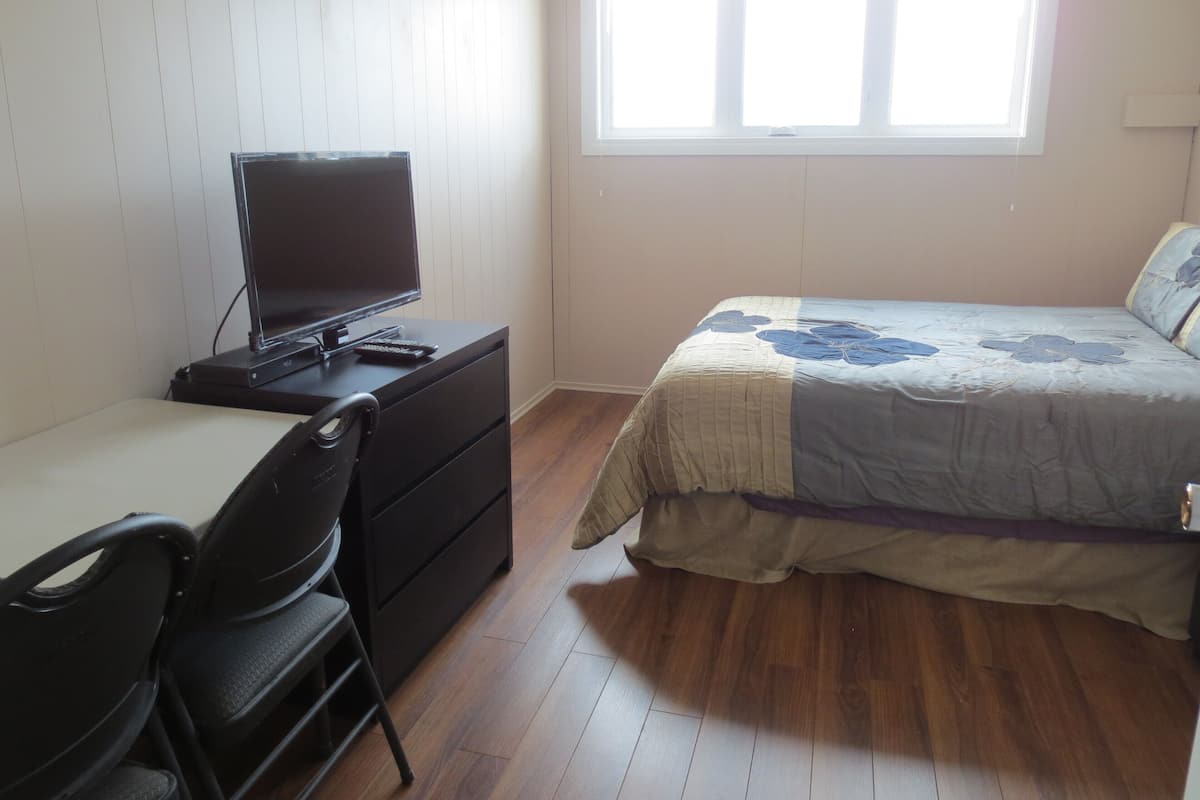 Quarto com cama, escrivaninha e computador, e uma janela.