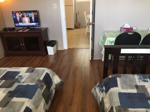 Ein Hotelzimmer mit zwei Betten, einem Fernseher, der eine Nachrichtensendung zeigt, einem Essbereich mit Tisch und Stühlen sowie einem Mülleimer.