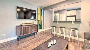 A modern living room with a dark wood floor, a central coffee table with candles, a mounted TV, and a kitchen area visible through an open doorway. - NEW! Centrally Located Charlotte Home by Amtrak! (Charlotte)