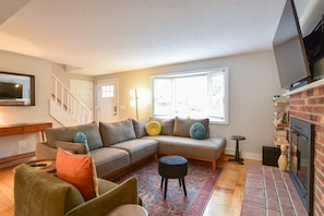 A modern living room with a sectional sofa, a small round ottoman, a fireplace, and a staircase leading to another level. - Sunroom Expansive Patio Fenced Yard! (Eastham)