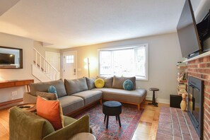 A living room with a sectional sofa, a small round ottoman, a TV mounted on the wall, a fireplace, and a staircase.