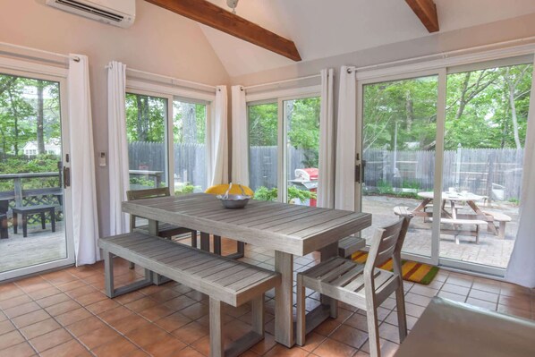 A patio area with a wooden table and benches, large sliding glass doors leading to a backyard with a barbecue grill and a wooden deck.