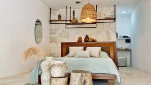 A modern bedroom with a stone wall, a wooden bed frame, a hanging light fixture, and a small kitchenette in the background. - Casa Macay (Tulum)