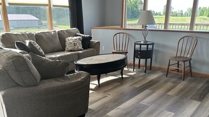 Un salon comprenant un canapé d’angle, une table basse ronde, deux chaises en bois et une lampe posée sur une petite table.