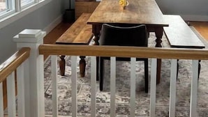 A dining area with a wooden table and chairs, a hanging light fixture, and a view of the outside through large windows.