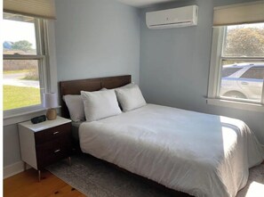 A bedroom with a bed, a nightstand, a lamp, and an air conditioning unit.