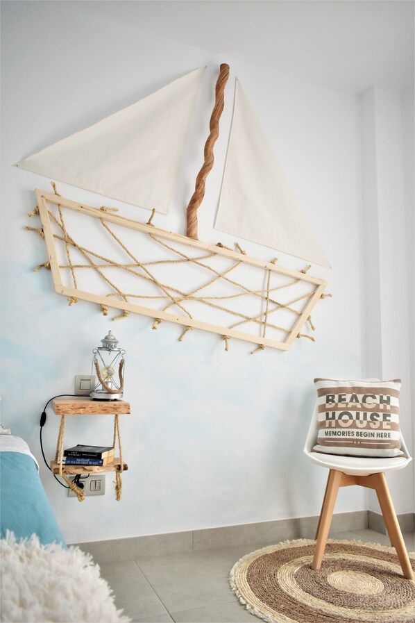 Una habitación con una decoración de velero montado en la pared, un pequeño estante de madera con libros y un farol, y una silla con un cojín que dice 'Beach House Memories Begin Here'.