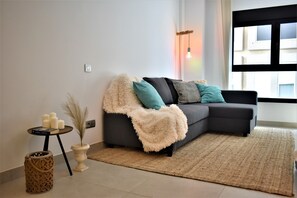 Una sala de estar moderna con un sofá gris oscuro, cojines azul turquesa y blancos, una manta blanca mullida, una mesita auxiliar con velas y una cesta tejida.