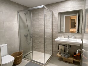 Un baño moderno con ducha de vidrio, lavamanos blanco y repisa de madera con toallas y artículos de aseo.