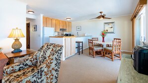 A compact living space with a kitchenette, a dining area with wooden chairs, a sofa with a leaf pattern, and a ceiling fan. - Ocean View 2 Bedroom Kahalu'u Reef KR301 (Kailua Kona)