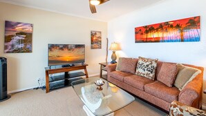 A living room with a ceiling fan, a flat-screen TV, a sofa, a coffee table, and wall art. - Ocean View 2 Bedroom Kahalu'u Reef KR301 (Kailua Kona)