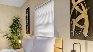 A bedroom with a bed, a wall-mounted light, a window with blinds, and a decorative wall piece.