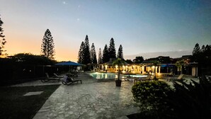 Área de piscina com espreguiçadeiras e guarda-sóis, cercada por árvores e edifícios.