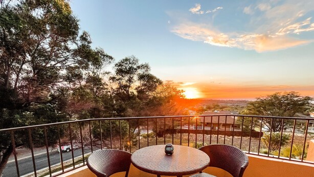 Varanda com mesa redonda e duas cadeiras, com vista para o pôr do sol, árvores e um edifício ao longe.