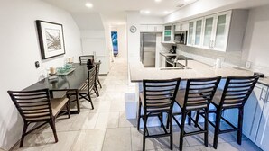 Uma cozinha moderna com armários brancos, uma área de refeições com uma mesa e cadeiras pretas, e um relógio visível na parede.