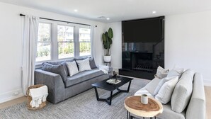 Ein modernes Wohnzimmer mit einem grauen Sofa, einem schwarzen Couchtisch, einem Kamin und einem großen Fenster mit Vorhängen.