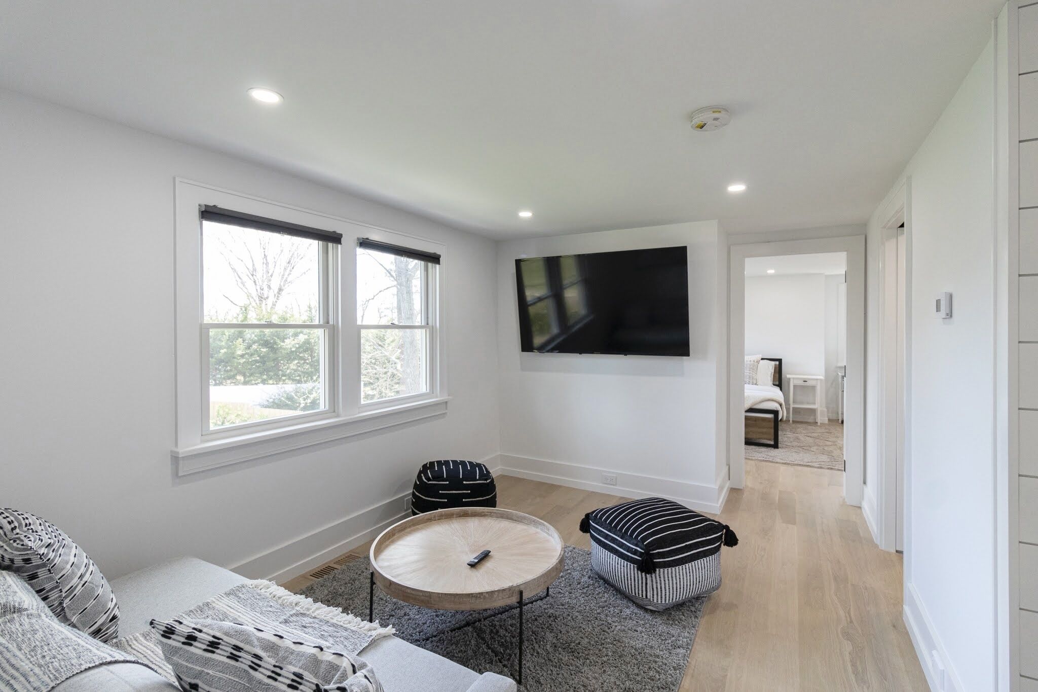 A modern living room with a large window, a flat-screen TV mounted on the wall, a round wooden coffee table, and a grey rug.