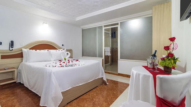 Habitación de hotel con cama, una mesita con un florero, una silla y un baño visible a través de una puerta abierta.