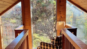 A cozy attic room with a checkered armchair, a small table, and a view of trees through a large window.