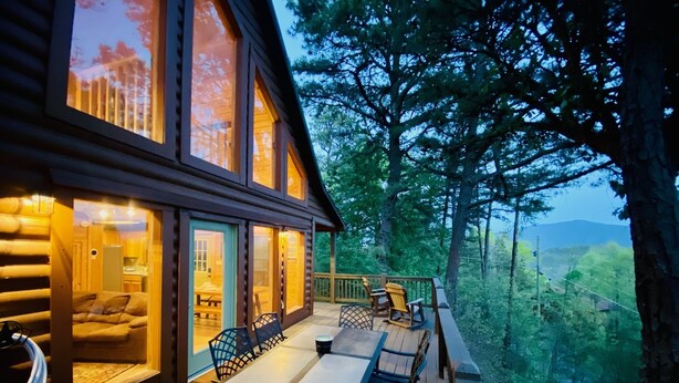 A log cabin with a deck, a dining area with chairs, and a view of the forest.