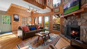 A cozy cabin interior with a stone fireplace, wooden walls, a leather sofa, and a large window overlooking a scenic view.